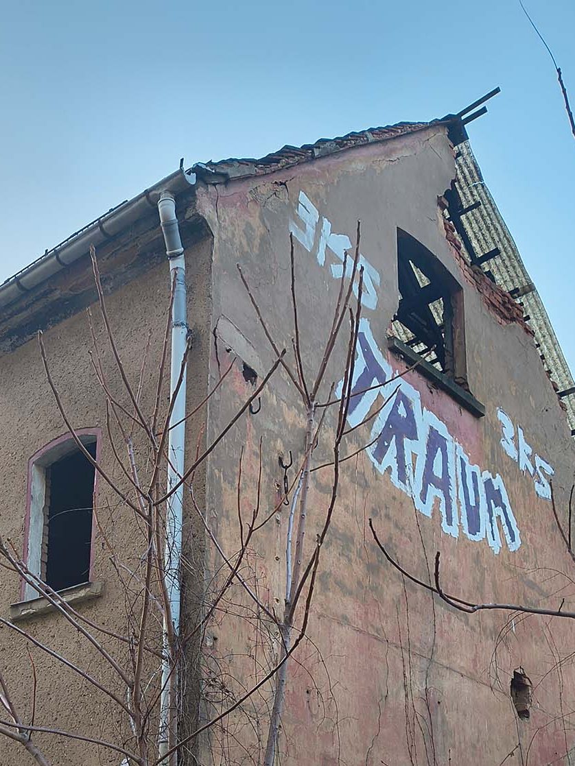 A close-up view of a dilapidated building with graffiti reading 'TRAUM' on its side. The structure shows signs of decay, with a broken window and peeling paint, surrounded by bare branches against a clear blue sky.
